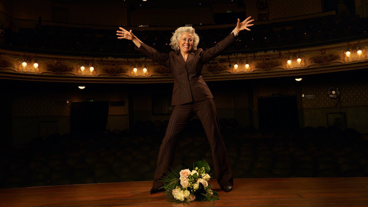 An image of Anna Longaretti on stage, wearing a black suit with her arms out. She stands above a bouquet of flowers.