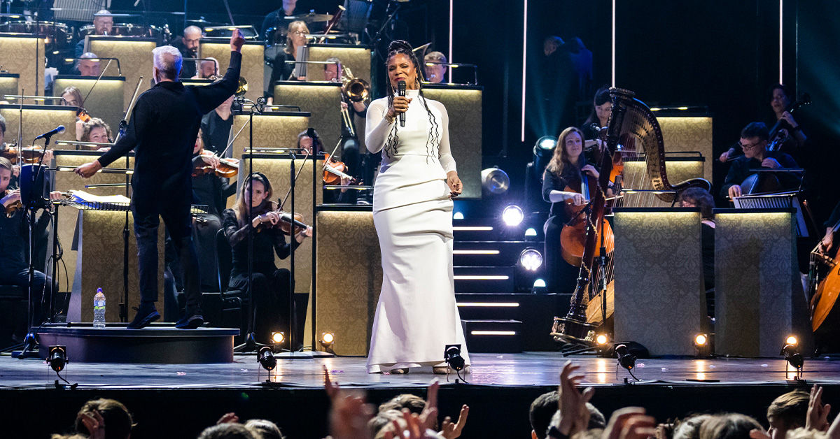 A black woman in white gown sings on stage, accompanied by an orchestra.
