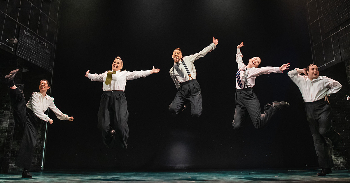 Five dancers captured mid-jump on stage, dressed in retro-inspired outfits with a simple black backdrop, expressing joy through dynamic leaps and open arms.